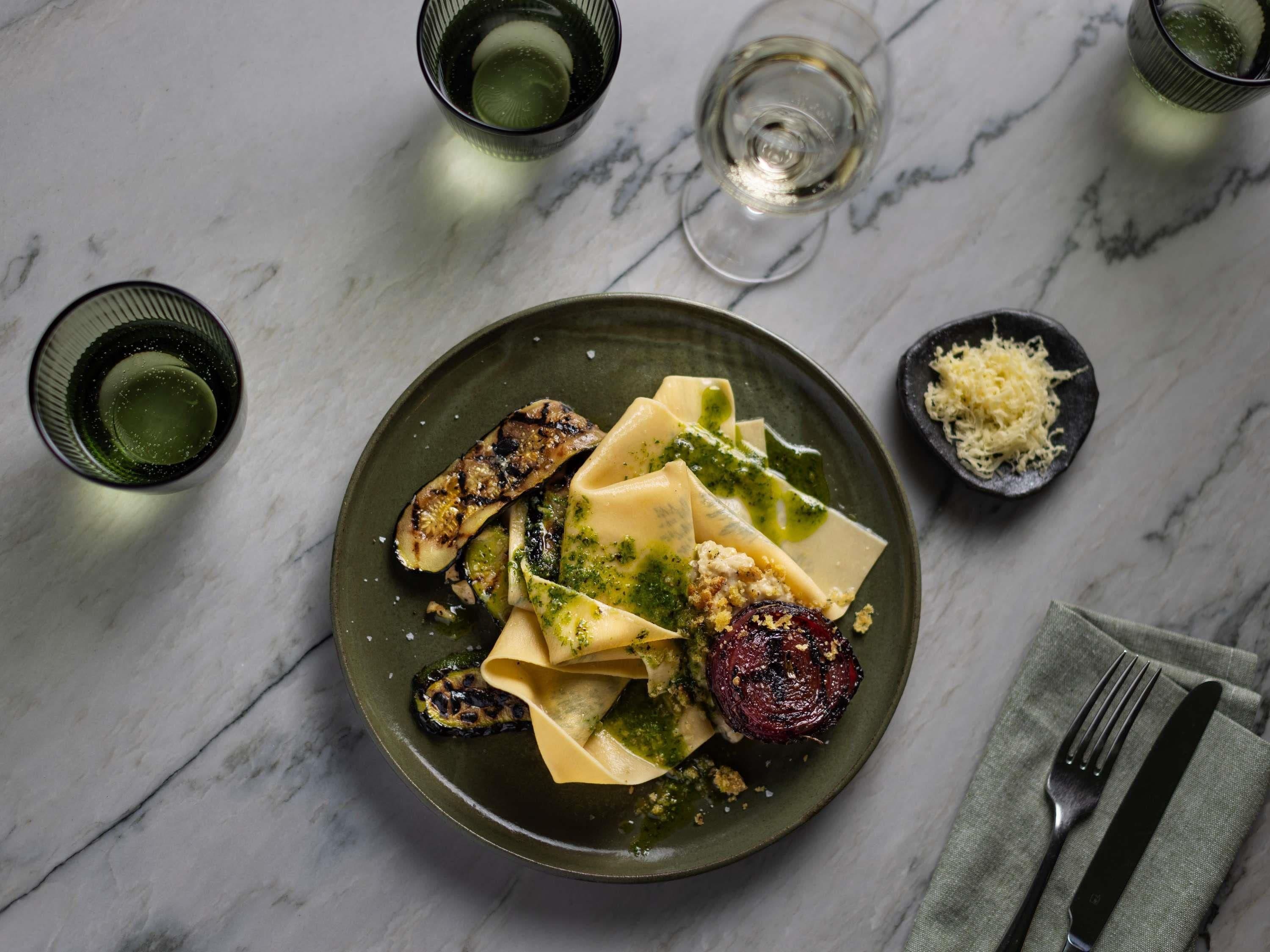Une assiette de pâtes, agrémentée de légumes et de fromage, est posée sur une table en marbre, accompagnée de verres et de couverts.