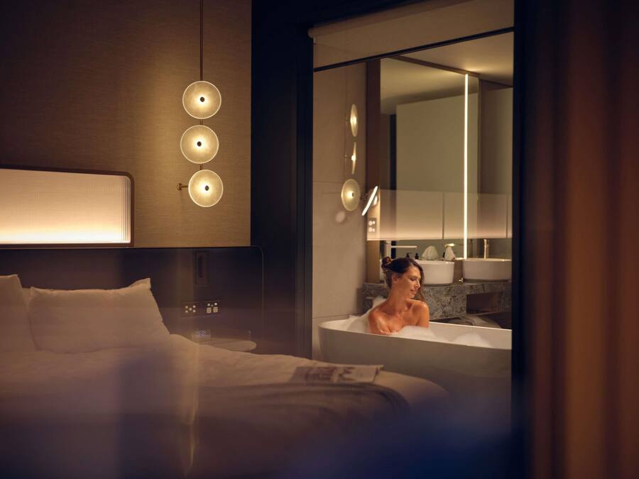 A hotel room with a bed, a nightstand, a wall-mounted light fixture, a bathroom with a bathtub, and a mirror.