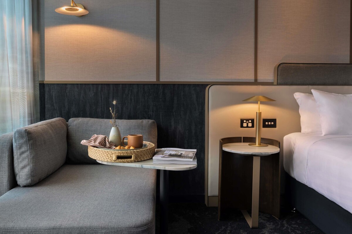A modern hotel room with a sofa, a small table with a basket, a lamp, and a bed.
