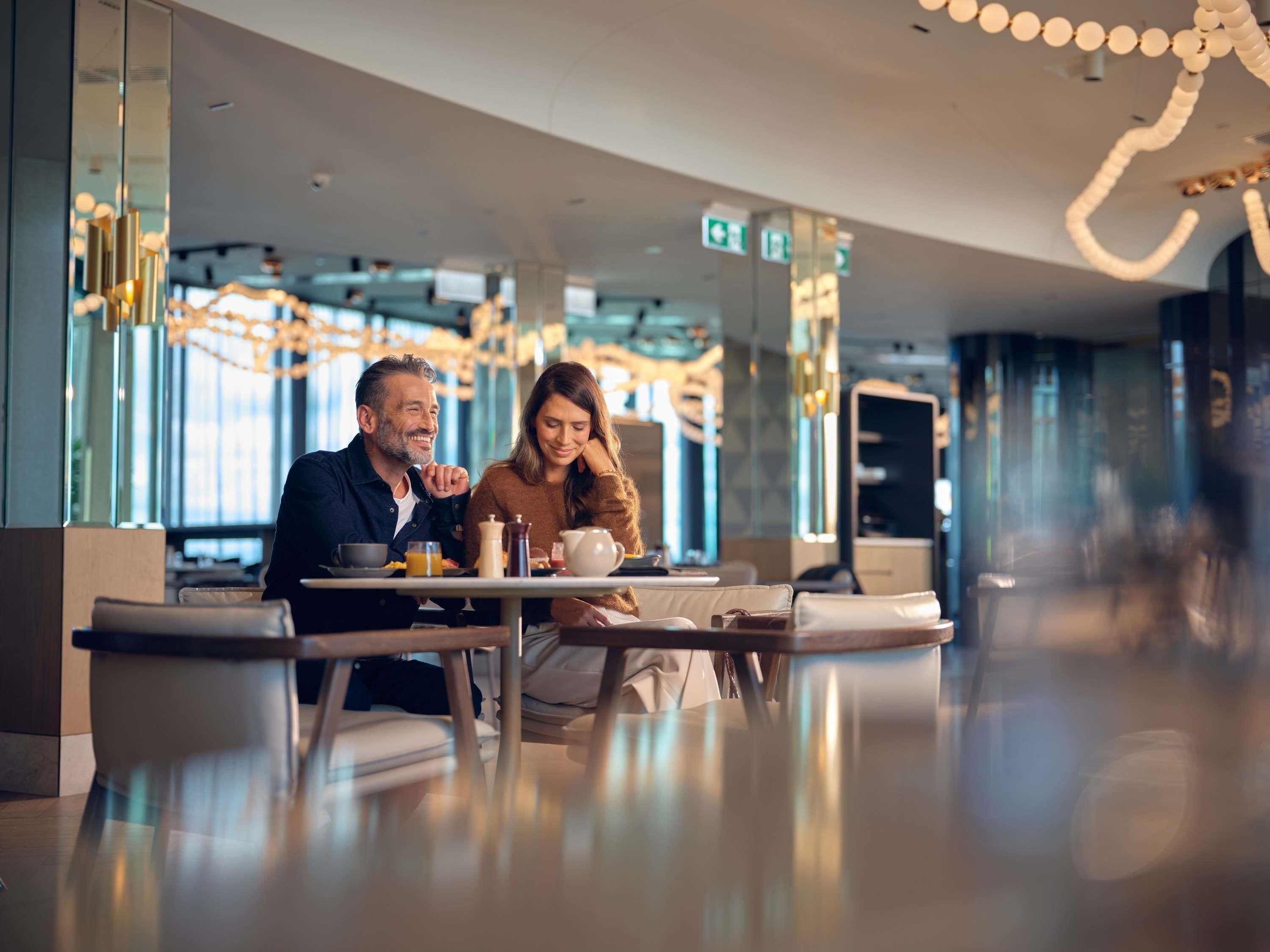 Un interior moderno con una pareja sentada a una mesa con alimentos para el desayuno, ventanas amplias e iluminación contemporánea.