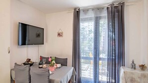 A dining area with a table set for two, a television mounted on the wall, and a view of the outdoors through a large window with curtains. - Holiday apartment close to the center (Njivice)