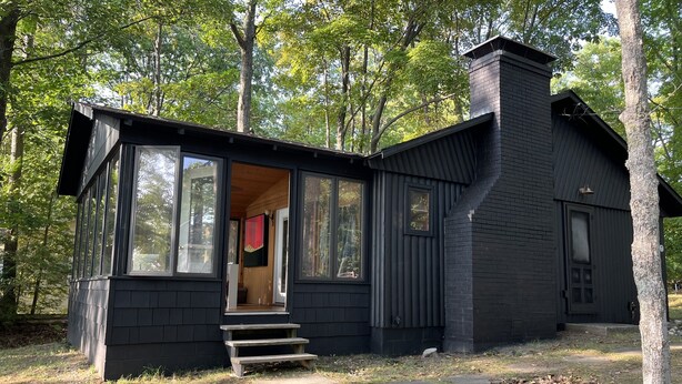 A black cabin with large windows, a chimney, and a wooden deck. - Lakefront Log Cabin with Contemporary touches (Carp Lake)