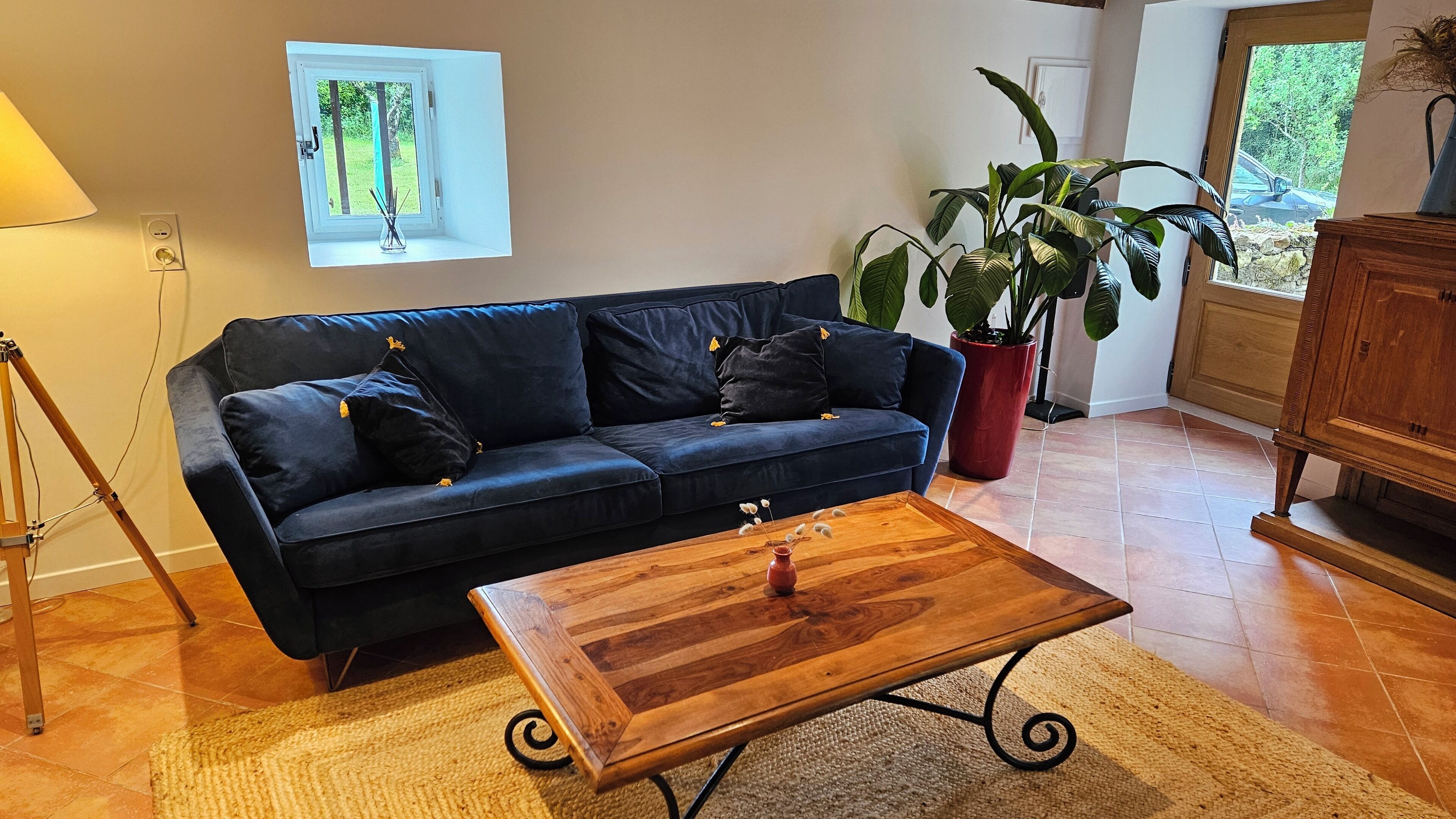 Un salon avec un canapé bleu foncé, une table basse en bois, une plante en pot et une fenêtre donnant sur un paysage verdoyant.