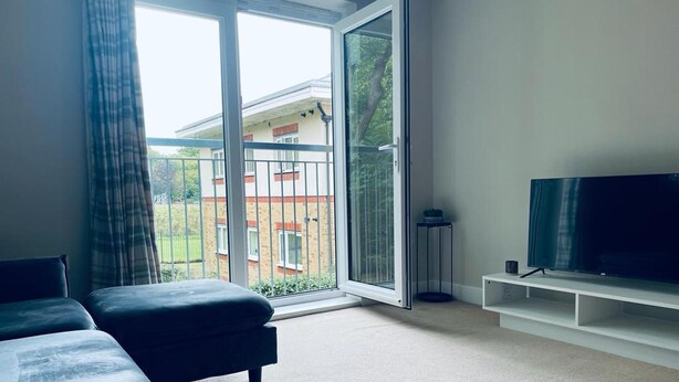 A modern living room with a large sliding glass door leading to a balcony, a flat-screen TV, and a sofa. - Stunning 2-bed Apartment in Crawley (Crawley)