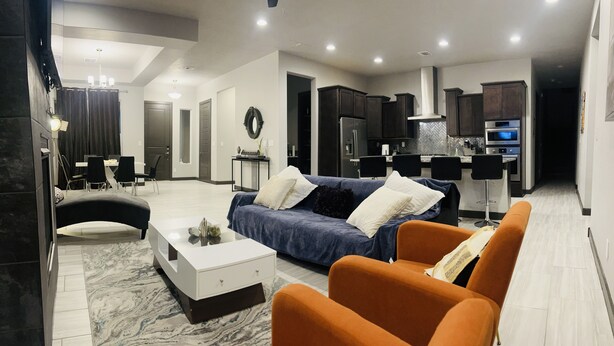 A modern living room with a sofa, armchairs, and a coffee table. There is a kitchen area with a refrigerator and microwave.