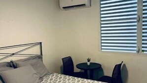 Une chambre à coucher comprenant un lit, un ventilateur de plafond, un climatiseur, une petite table avec des chaises et une fenêtre avec des stores.