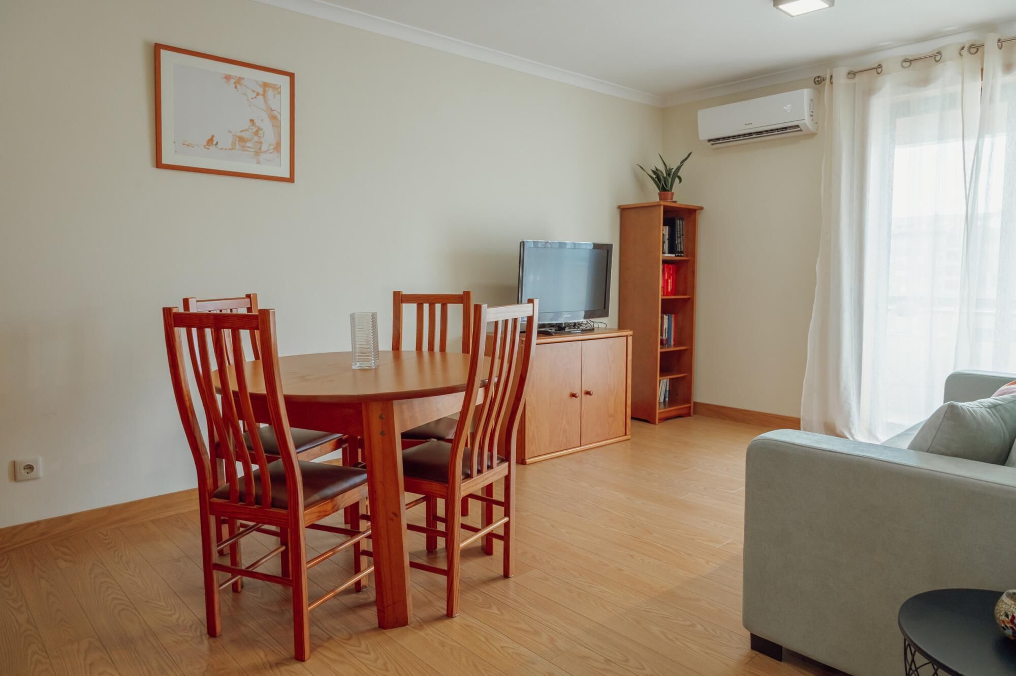 A living room with a dining table, chairs, a sofa, a TV, a bookshelf, and an air conditioning unit.