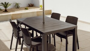 Una mesa rectangular al aire libre con seis sillas, cada una con asiento y respaldo tejido. La mesa se sostiene con un pilar central.
