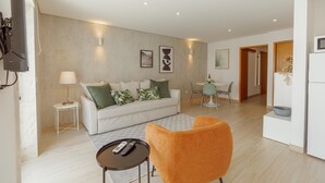Una sala moderna con un sofá, una mesa de centro y un sillón naranja brillante. Hay una zona de comedor con una mesa y sillas, y una cocina con un refrigerador.