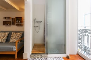 A room with a glass shower door, a wooden bench, a patterned rug, and a sofa with floral cushions. - Santos Tiny Studio by Homing (Lisbon)