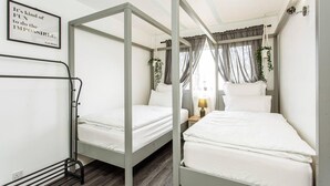 Una habitación con dos literas, una mesita de noche con lámpara, una cita enmarcada en la pared y una ventana con cortinas.