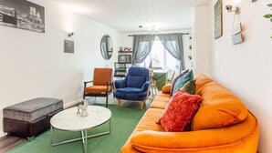 Una sala de estar con un sofá, dos sillones, una mesa de centro y un pequeño reposapiés. En el fondo, se observa una zona de comedor con una mesa y sillas.