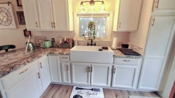 Una cocina con armarios blancos, un lavamanos, una encimera y un ventanal con cortinas.