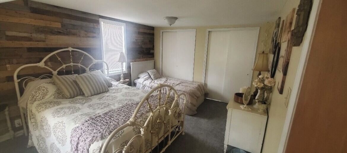 A bedroom with two beds, wooden paneling on the walls, a window with blinds, and a dresser with decorative items.