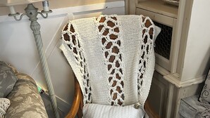 Una silla de madera con un plaid de ganchillo y un cojín, colocada junto a una lámpara y un almohadón estampado.