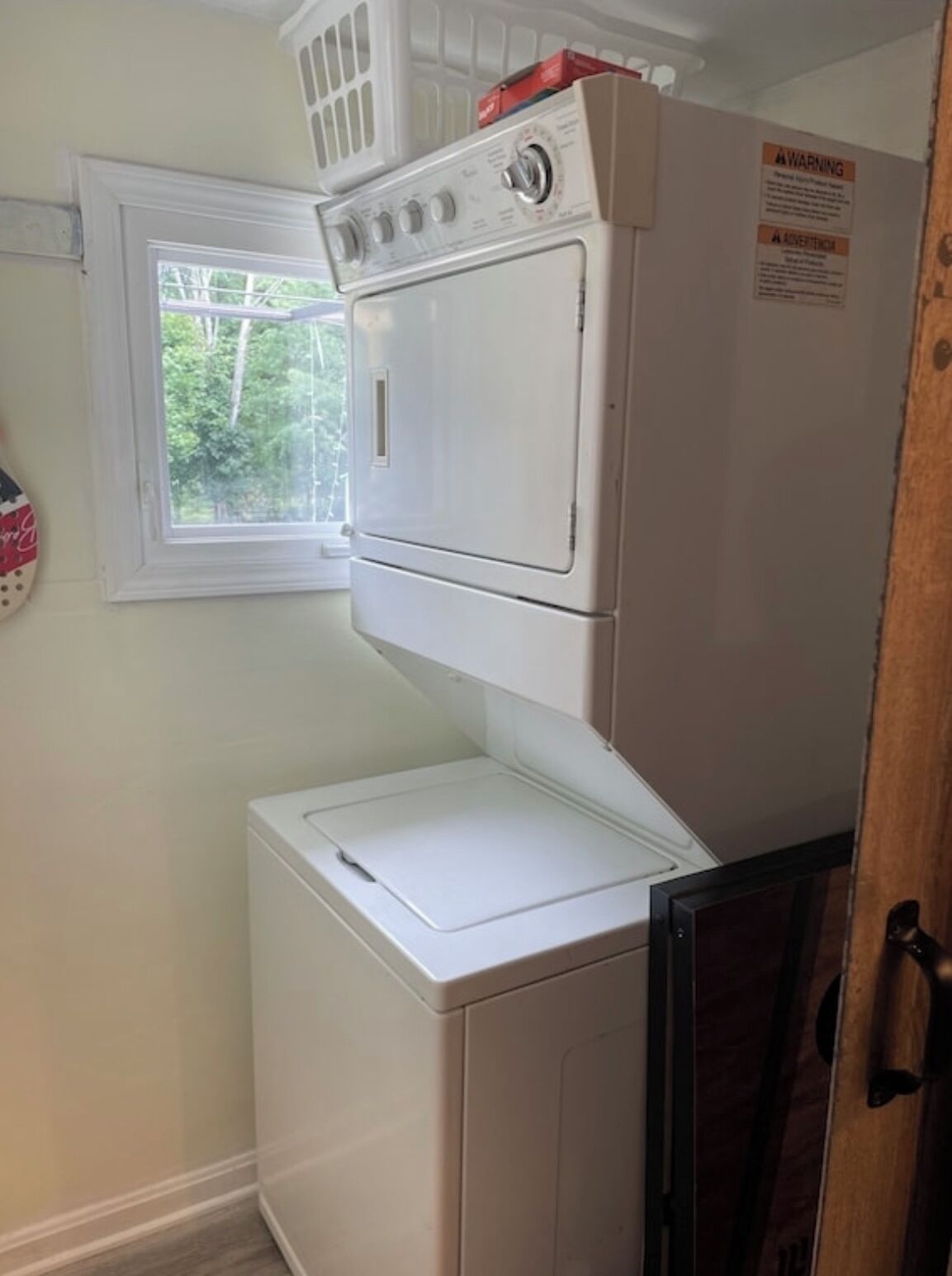 Une buanderie équipée d’une machine à laver et d’un sèche-linge superposés, une fenêtre donnant sur un espace verdoyant et une porte dotée d’une poignée.