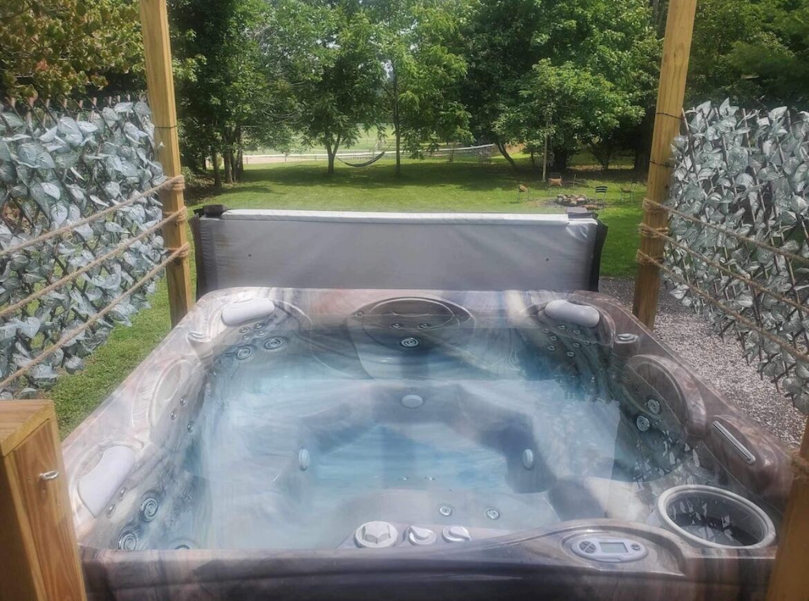 A hot tub with a wooden frame, surrounded by a stone wall, set in a garden with trees and a hammock in the background.