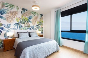 A bedroom with a large bed, a nightstand, a wall with a tropical leaf pattern, and a view of the sea through a window. - Las Caletillas Sea View Penthouse (Candelaria)