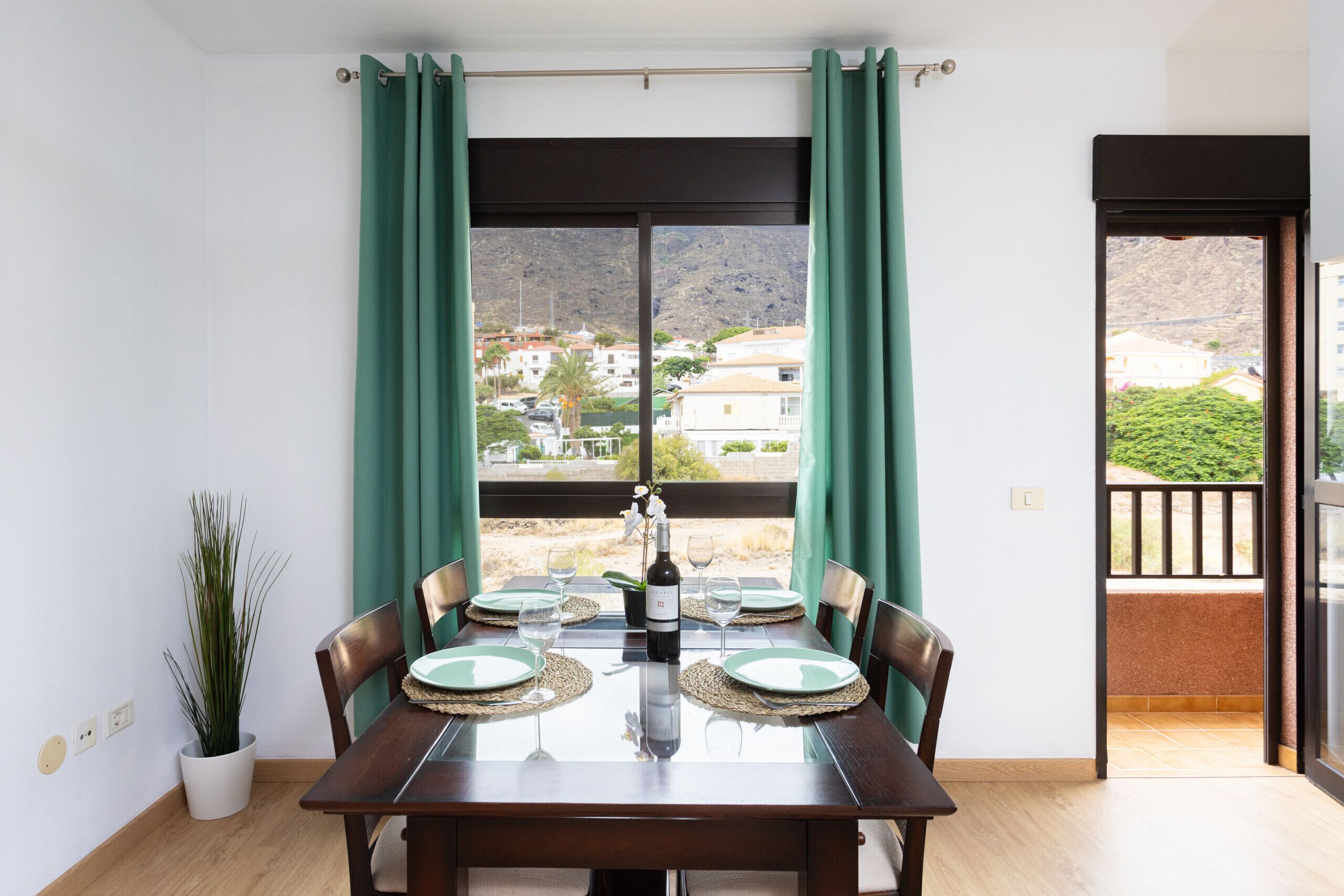 A dining area with a table set for four, a potted plant, and a view of a mountainous landscape through large windows.