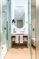 A modern bathroom with a glass shower enclosure, a marble-tiled wall, a white sink with a large mirror, and a wooden floor.