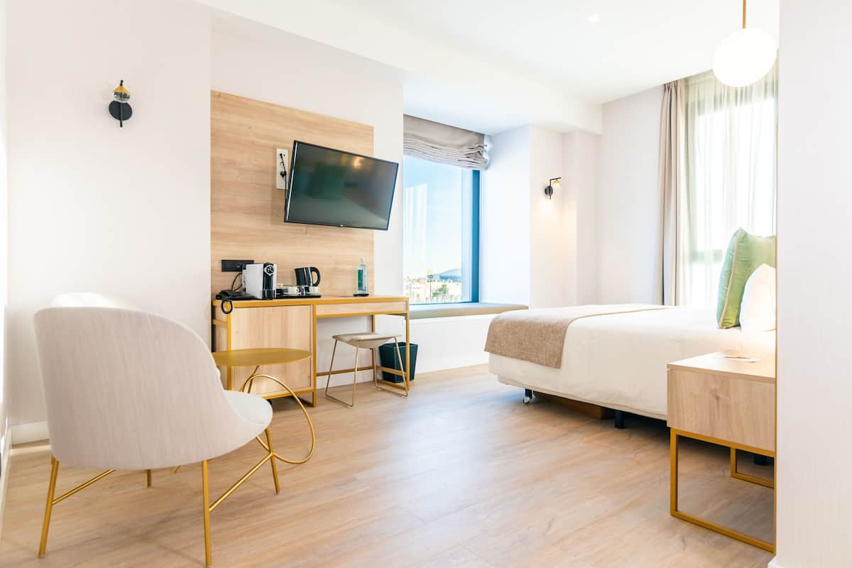 A modern hotel room with a bed, desk, chair, TV, and a view of the city.