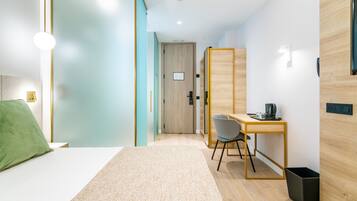 Una habitación de hotel moderna con una cama, un escritorio y una silla. Se observa una puerta que conduce a otra habitación y un cubo de basura.