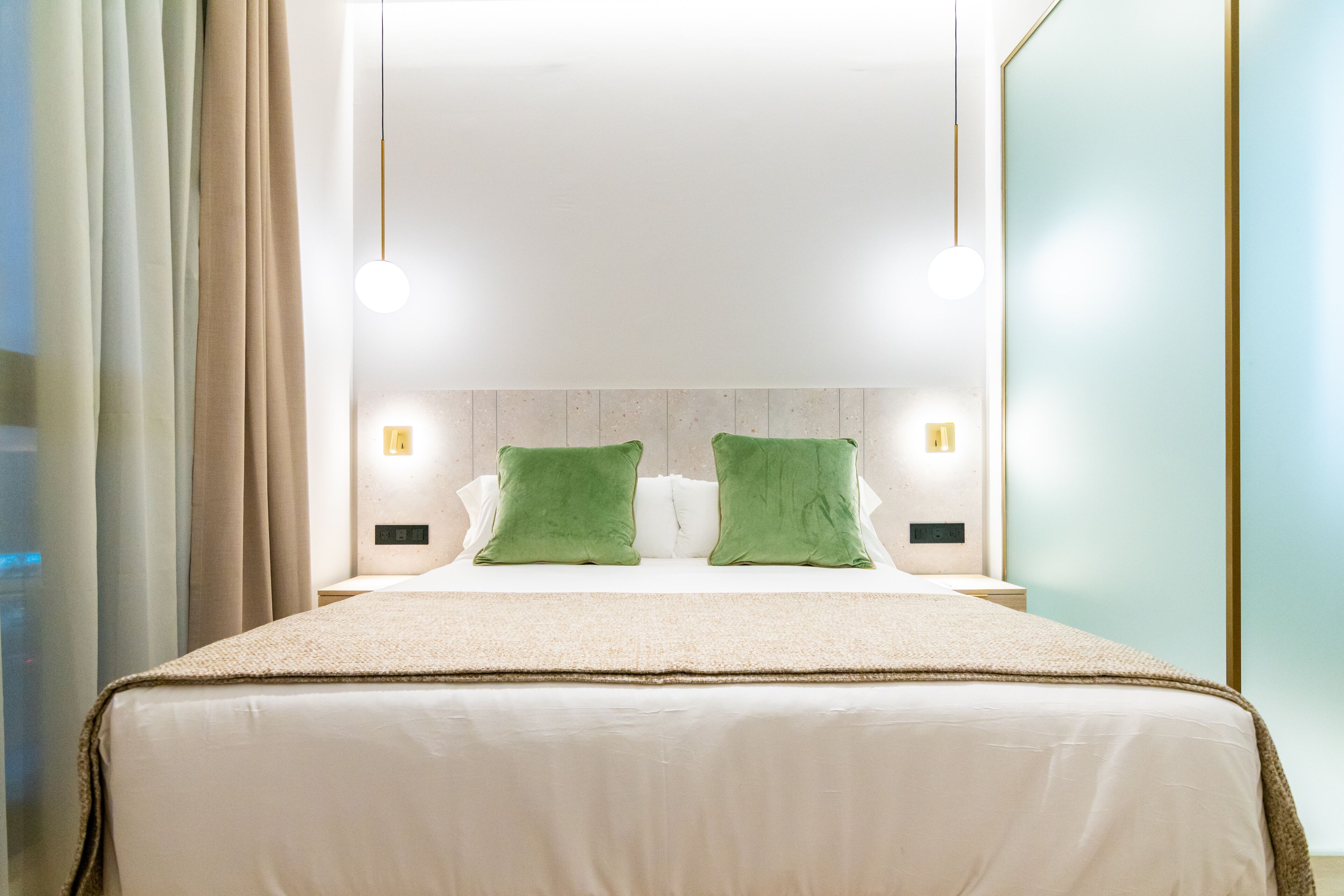 A neatly made bed with a beige bedspread and two green pillows, a bedside table with a lamp, and a wall-mounted light fixture above the bed.