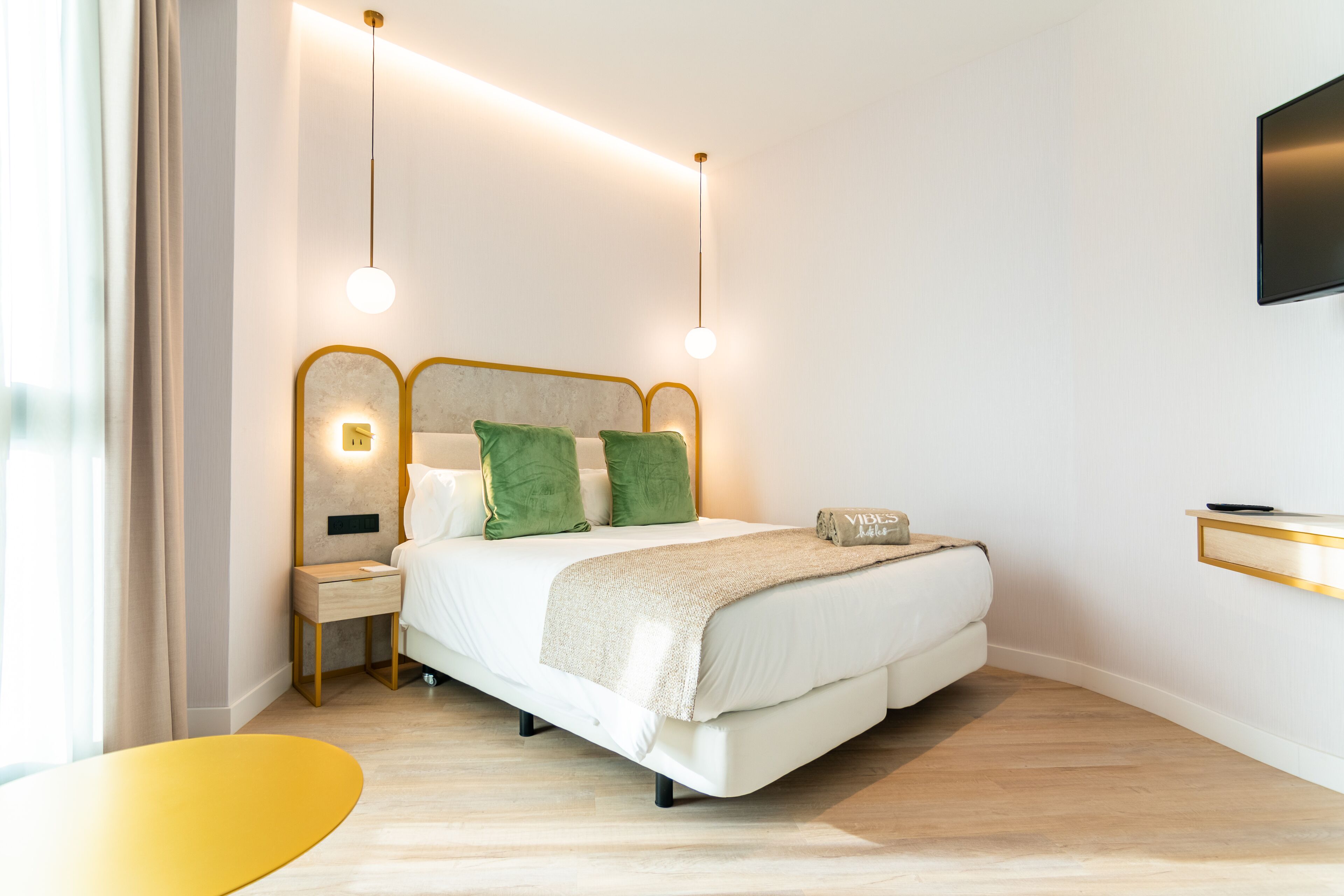Une chambre moderne avec un lit, des tables de chevet, une télévision et un fauteuil jaune.