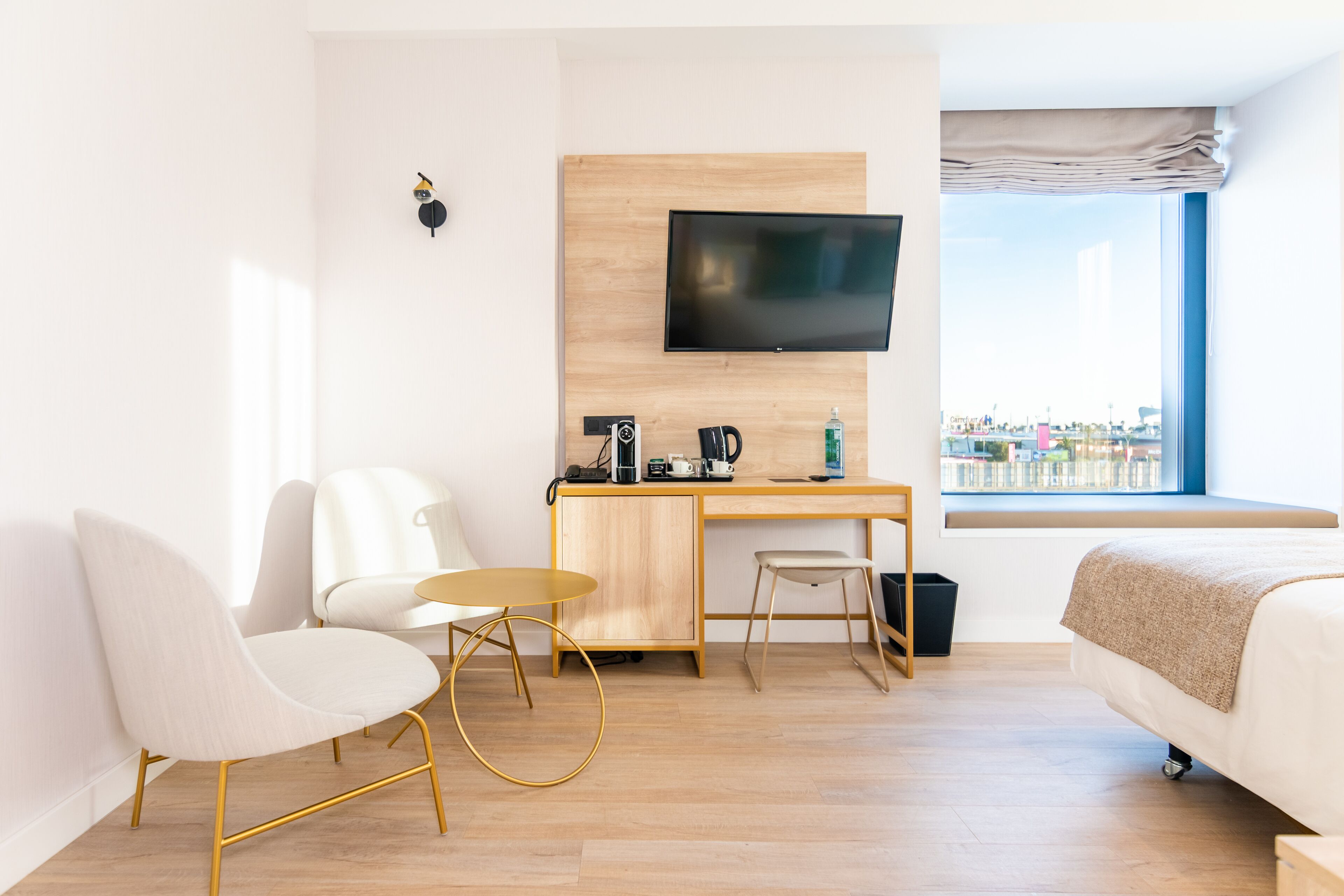 A modern hotel room with a bed, a desk with a TV, a chair, and a view of the city.