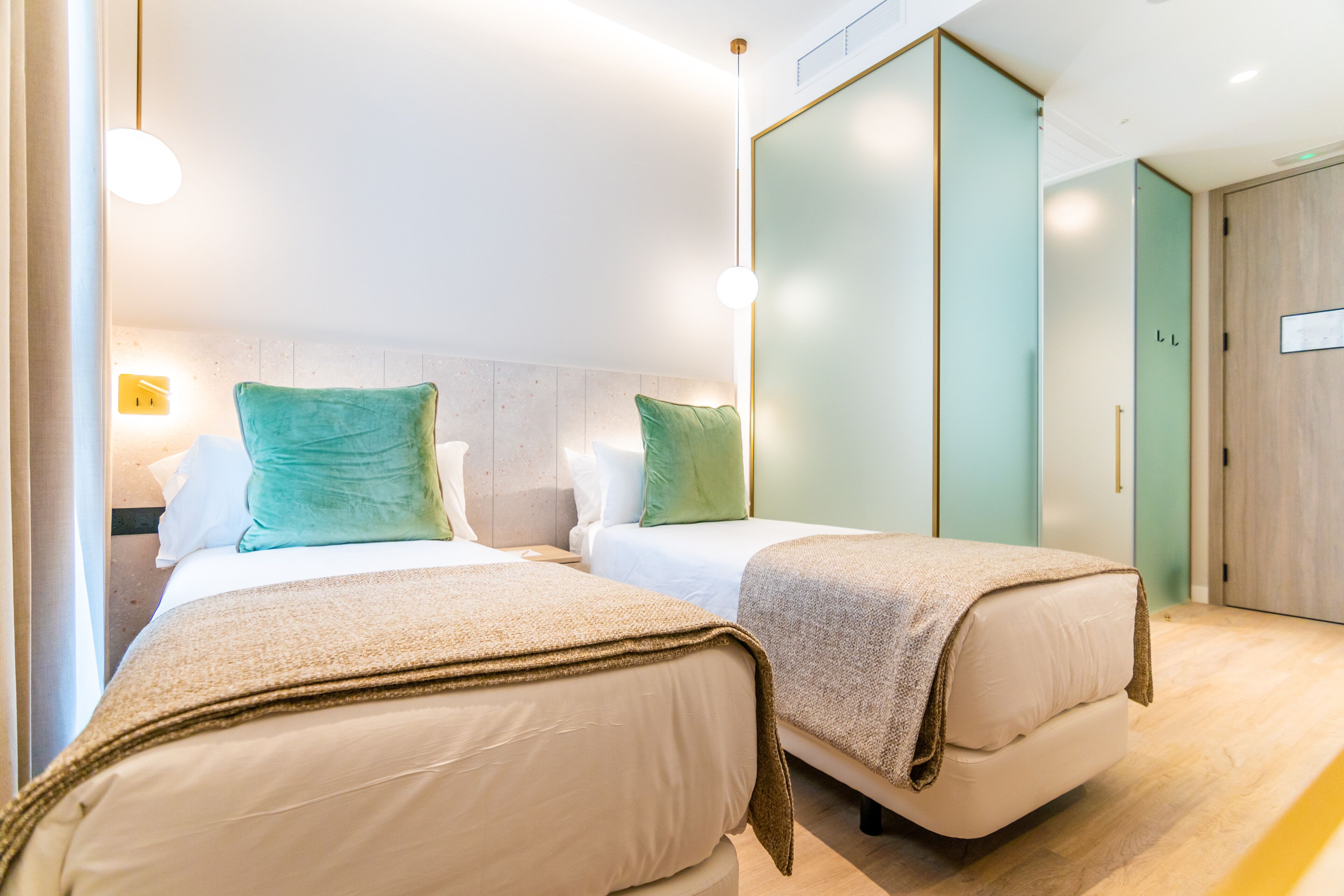 A hotel room with two beds, each with a green pillow and a beige blanket. There is a wooden wardrobe and a door with a sign.