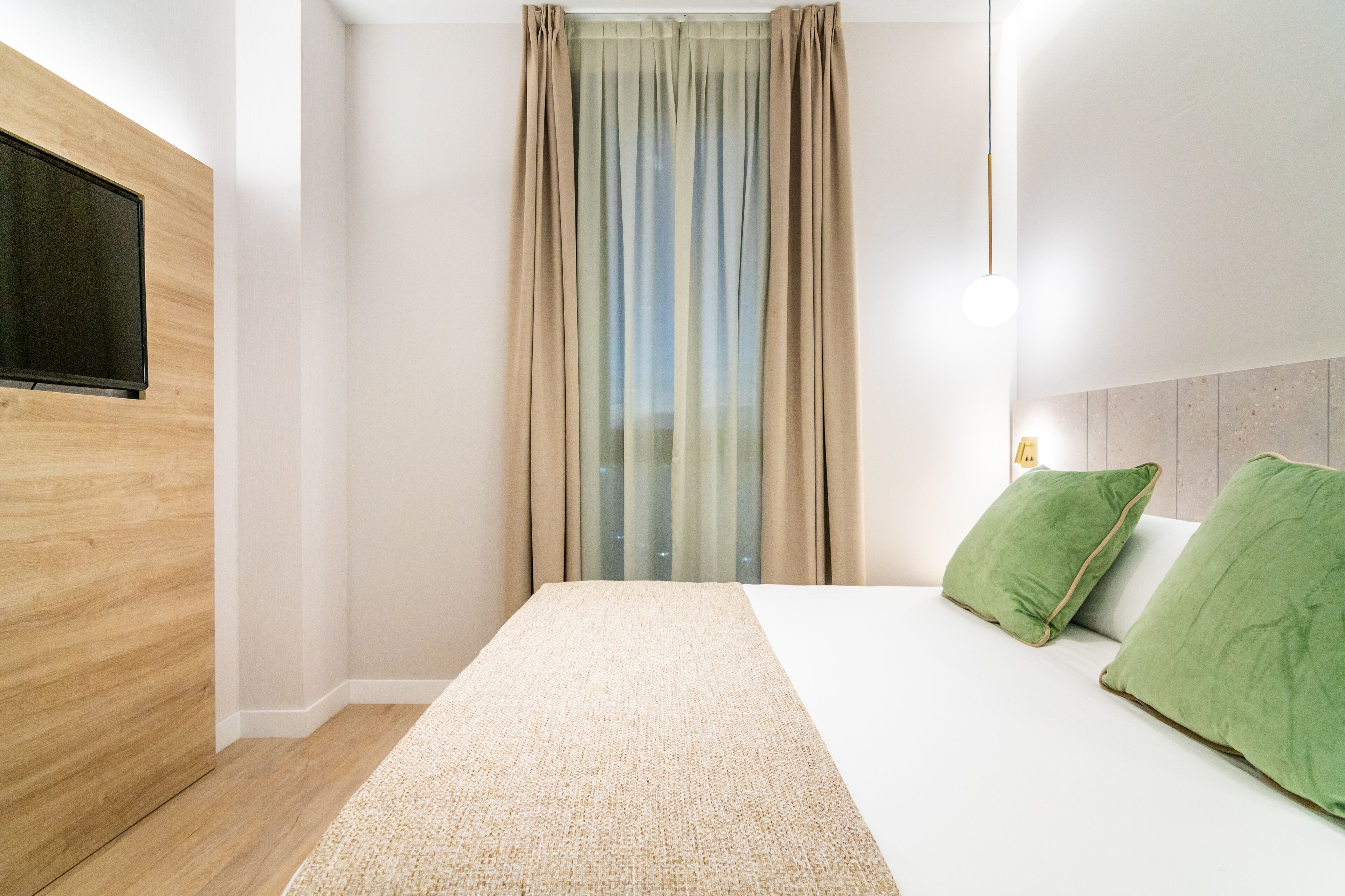 Ein modernes Hotelzimmer mit einer Wand in Holzoptik, einem großen Bett mit grünen Kissen, einem flachbildfernseher an der Wand und einem Fenster mit transparenten Vorhängen.