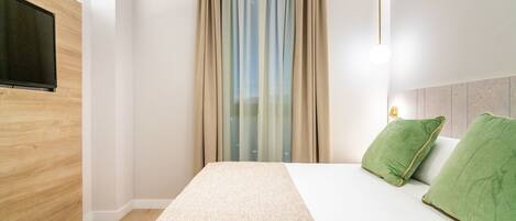 Quarto de hotel moderno com parede de destaque em madeira, uma cama grande com almofadas verdes, uma televisão de tela plana na parede e uma janela com cortinas leves.
