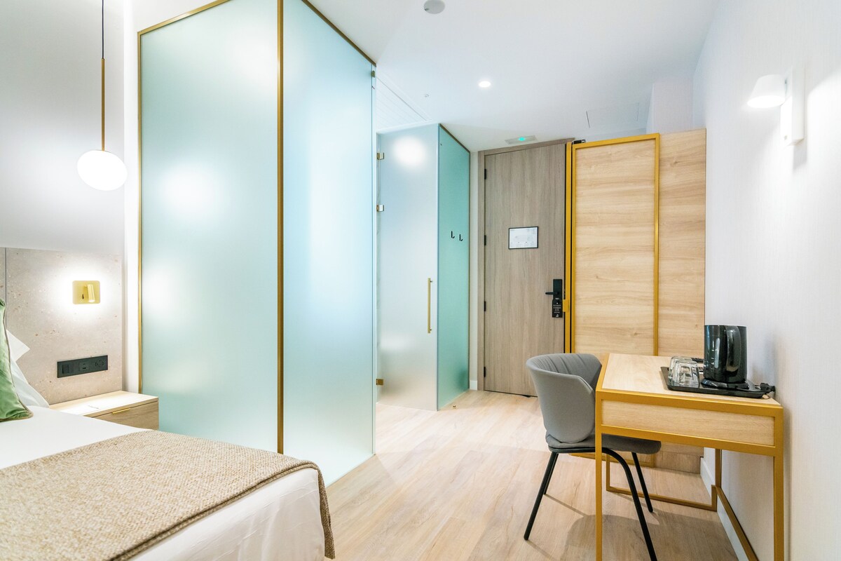 Une chambre d’hôtel moderne équipée d’un lit, d’un bureau avec une chaise, d’une machine à café et d’une porte bleu clair.