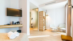 A modern hotel room with a flat-screen TV, a desk with a coffee maker, a sofa with towels, a wooden partition, and a seating area with chairs and a small table. - Hotel Málaga Vibes (Málaga)