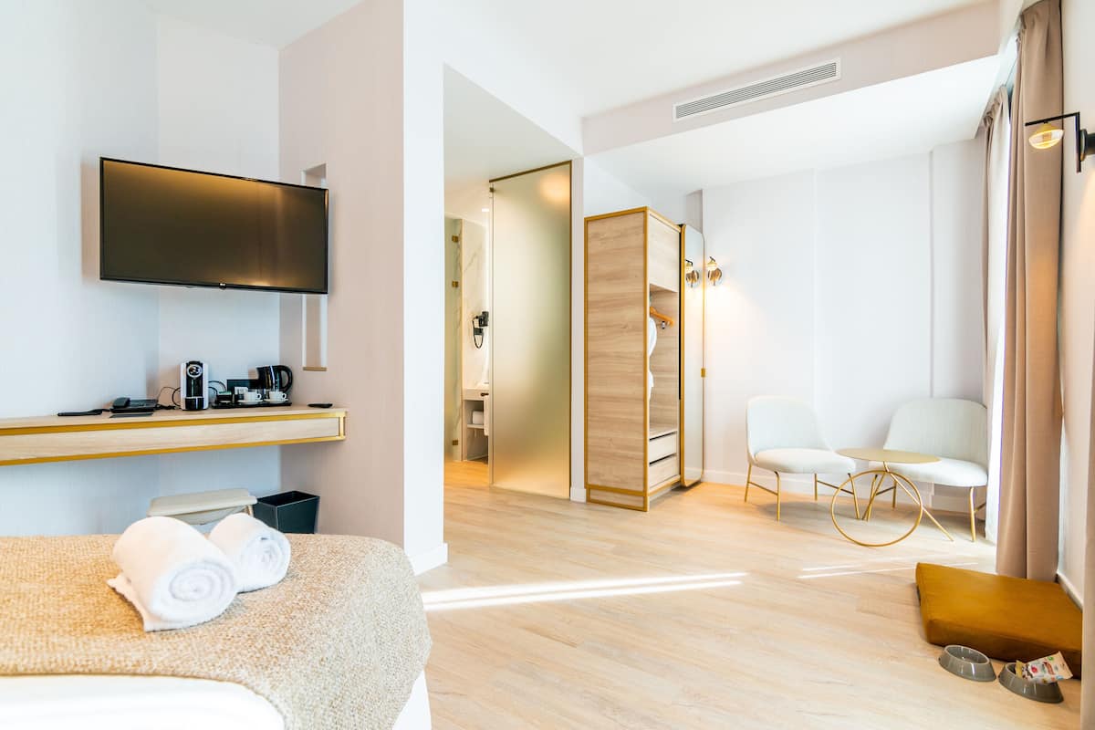 A modern hotel room with a flat-screen TV, a desk with a coffee maker, a sofa with towels, a wooden partition, and a seating area with chairs and a small table.