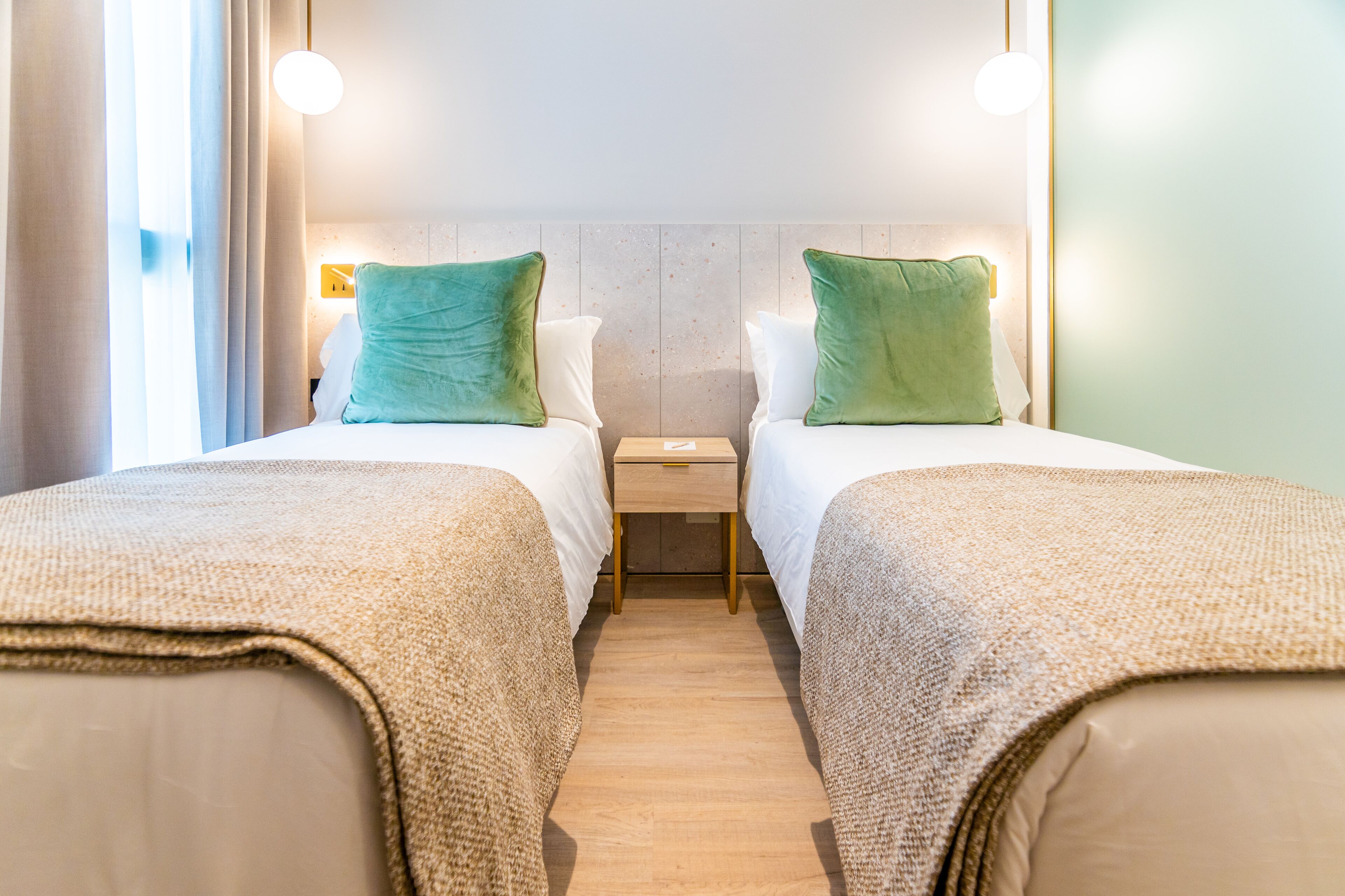 Camera d'albergo con due letti, ognuno con un cuscino verde e un comodino in legno a lato.