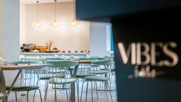 A modern dining area with light blue chairs and tables, a kitchen in the background, and hanging pendant lights.