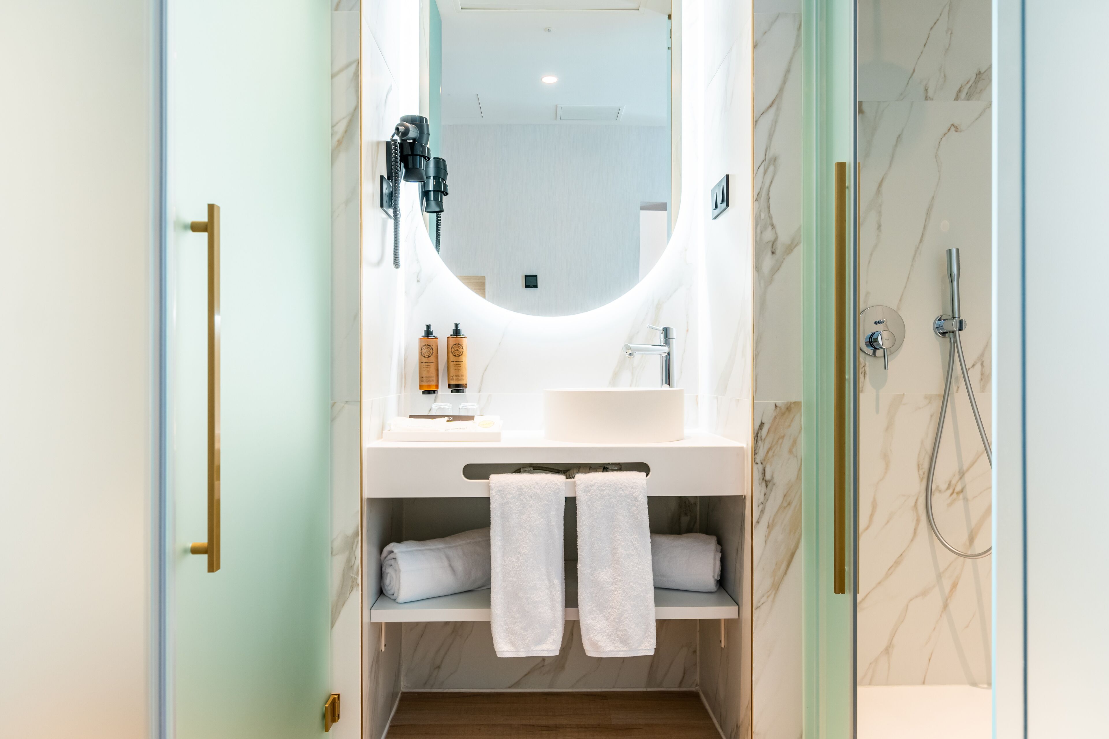 A bathroom with a large mirror, a sink, and a shower. There are towels hanging on a rack and toiletries on the sink.