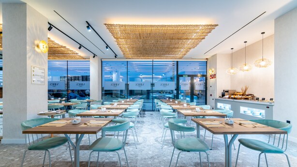 A modern restaurant with a central open kitchen, glass partitions, and a mix of metal and upholstered chairs. - Hotel Málaga Vibes (Málaga)
