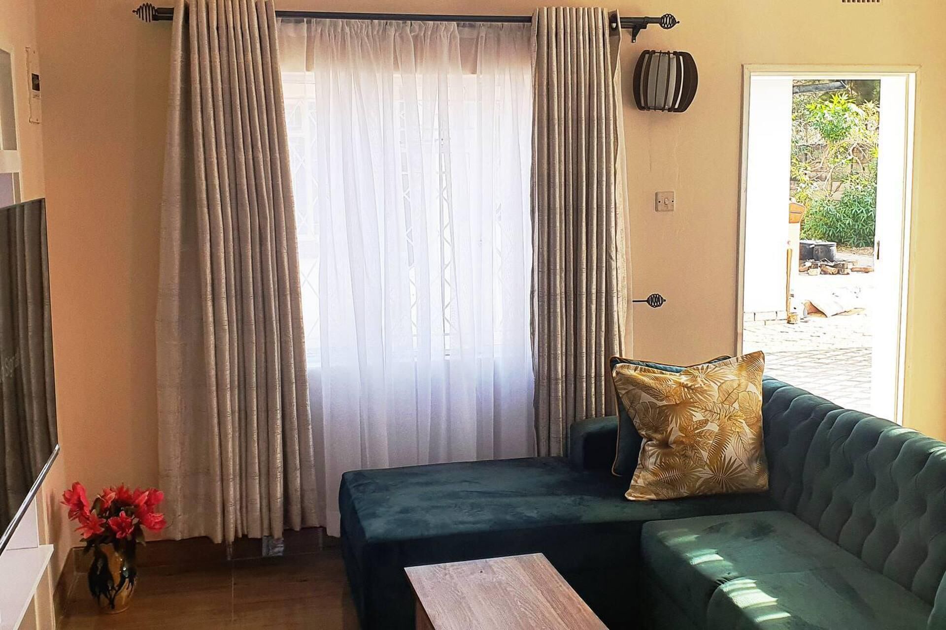 Ein Zimmer mit einem grünen Sofa, einem kleinen Tisch, einer Vase mit roten Blumen und Blick auf Grünfläche draußen.