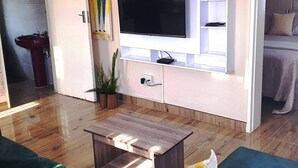 A modern living room with a flat-screen TV mounted on a white wall, wooden flooring, a small coffee table, and a potted plant. - Gorgeous 1-bed Apartment in Harare (Harare)