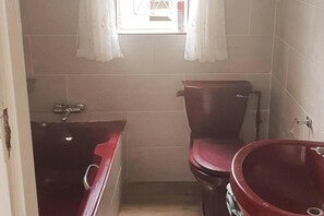 A small bathroom with a red sink, a toilet, and a window with white curtains.