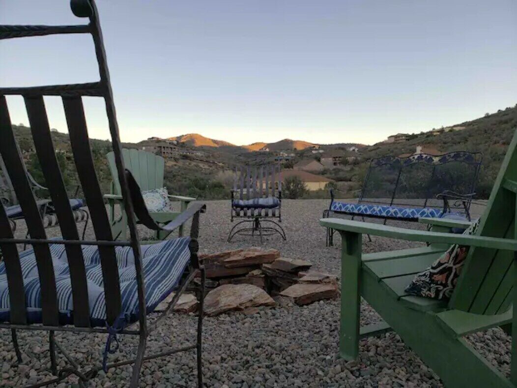 An outdoor seating area with chairs and a table, surrounded by gravel and rocks, overlooking a hilly landscape with buildings in the distance.