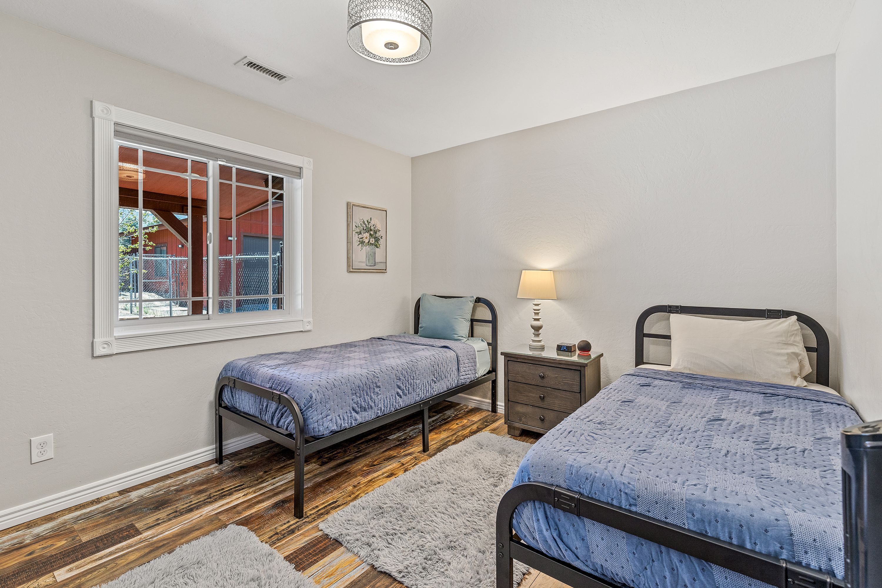 A bedroom with two single beds, a nightstand, a lamp, and a window with a view of a red building.