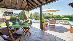 An outdoor patio with a covered seating area, a dining table set with plates and glasses, and a pool in the background. - Your vacation surrounded by greenery at Villa Irde (6 rooms 6 bathrooms) (Alghero)