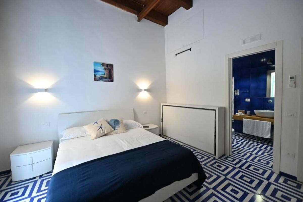 A bedroom with a bed, bedside tables, and a nightstand. There is a door leading to a bathroom with a blue tiled floor.