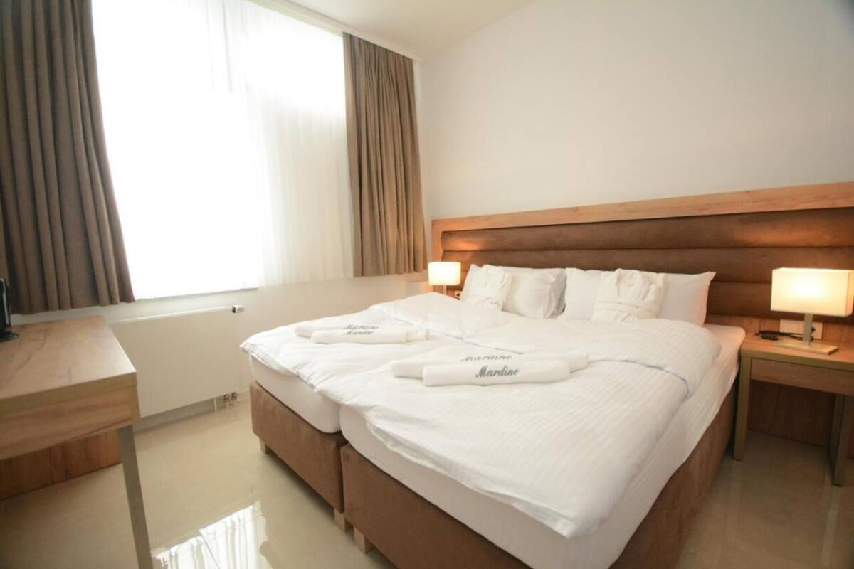 A hotel room with a large bed, two bedside tables, a wooden headboard, and a window with curtains.