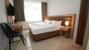 A hotel room with a wooden bed, a desk with a chair, a television mounted on the wall, and a window with curtains.
