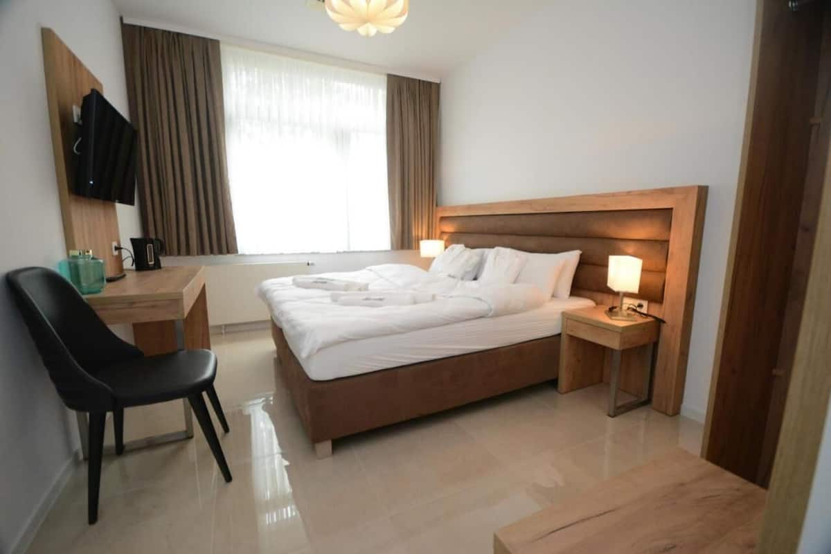 A hotel room with a wooden bed, a desk with a chair, a television mounted on the wall, and a window with curtains.