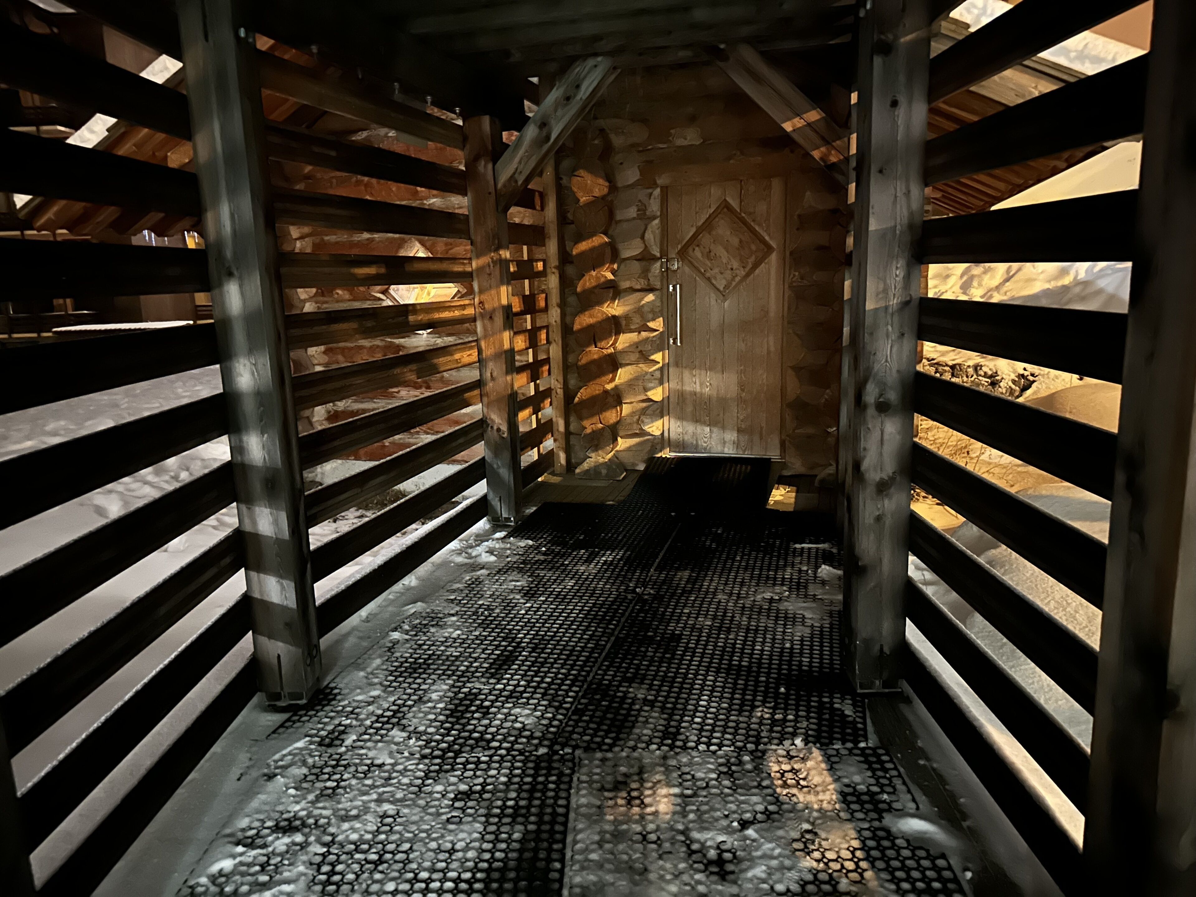 A narrow passageway with metal railings and a patterned floor, leading to a wooden door with a diamond-shaped design.
